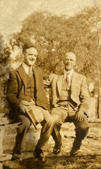 Two men sitting on a bench in front of trees.